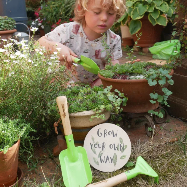 Un bambino sorridente che usa un set di attrezzi da giardinaggio in legno e metallo in un giardino.